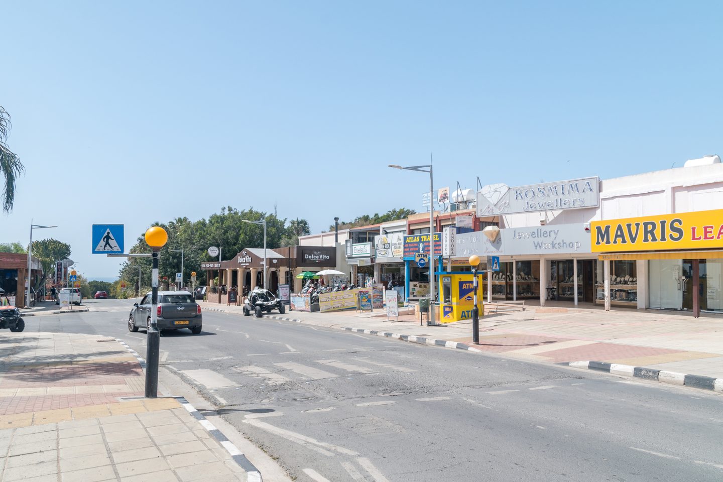 A sunlit resort shopping street of low shop units, among them a jeweller, a travel agent and a cash machine, with parked cars at the kerb and the sea visible downhill at the end of the road