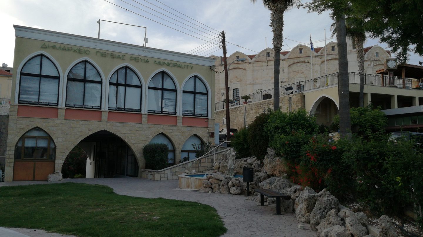 The arched stone municipality building on the square at Peyia, with palms and a flag above the terrace
