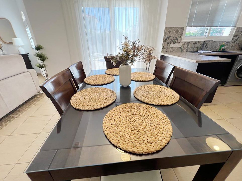Glass dining table set for six with woven placemats, between lounge and kitchen