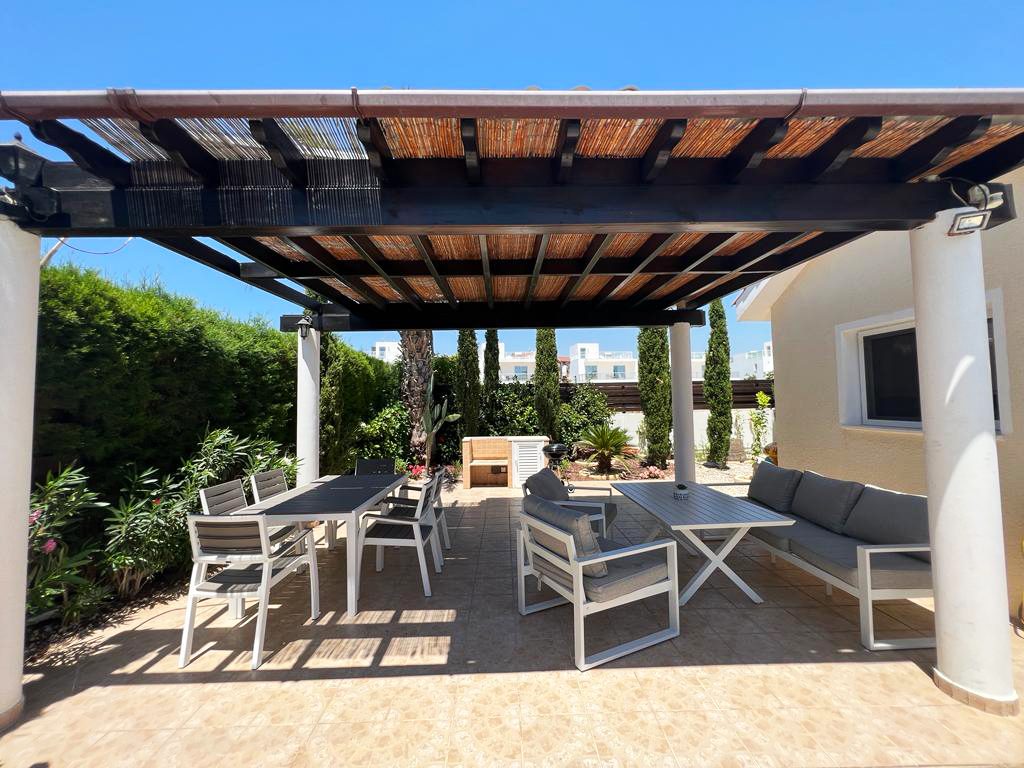 Long outdoor dining table and lounge seating beneath the pergola beside the barbecue