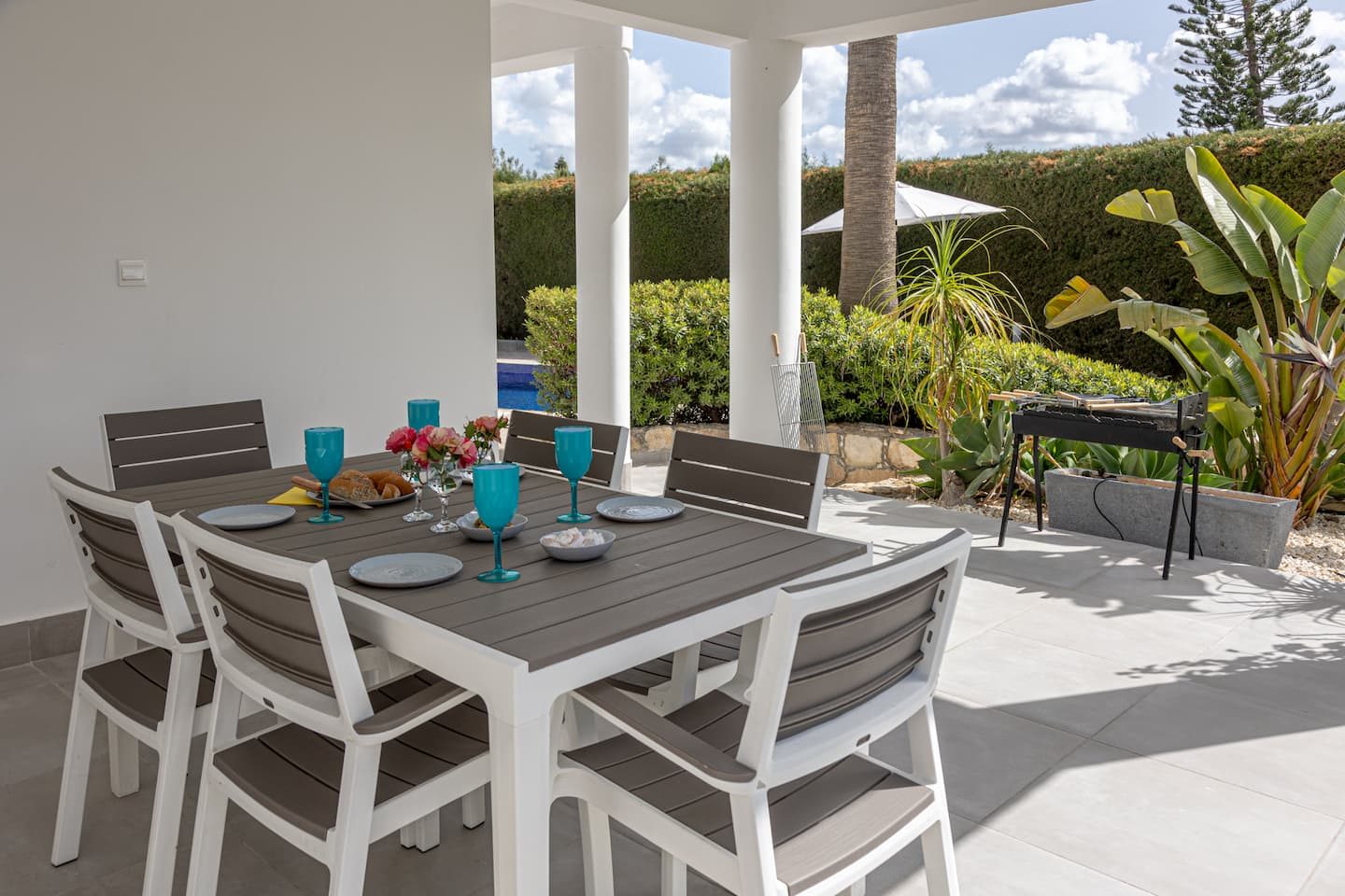 Outdoor table laid for six under the covered terrace, barbecue and garden beyond
