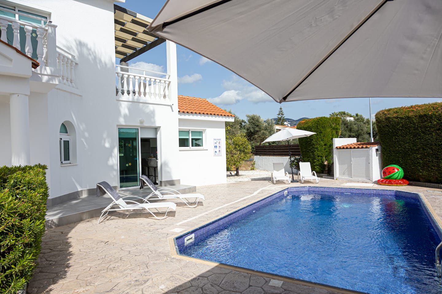 Shaded poolside terrace with parasol, white sun loungers and a rectangular pool beside the villa