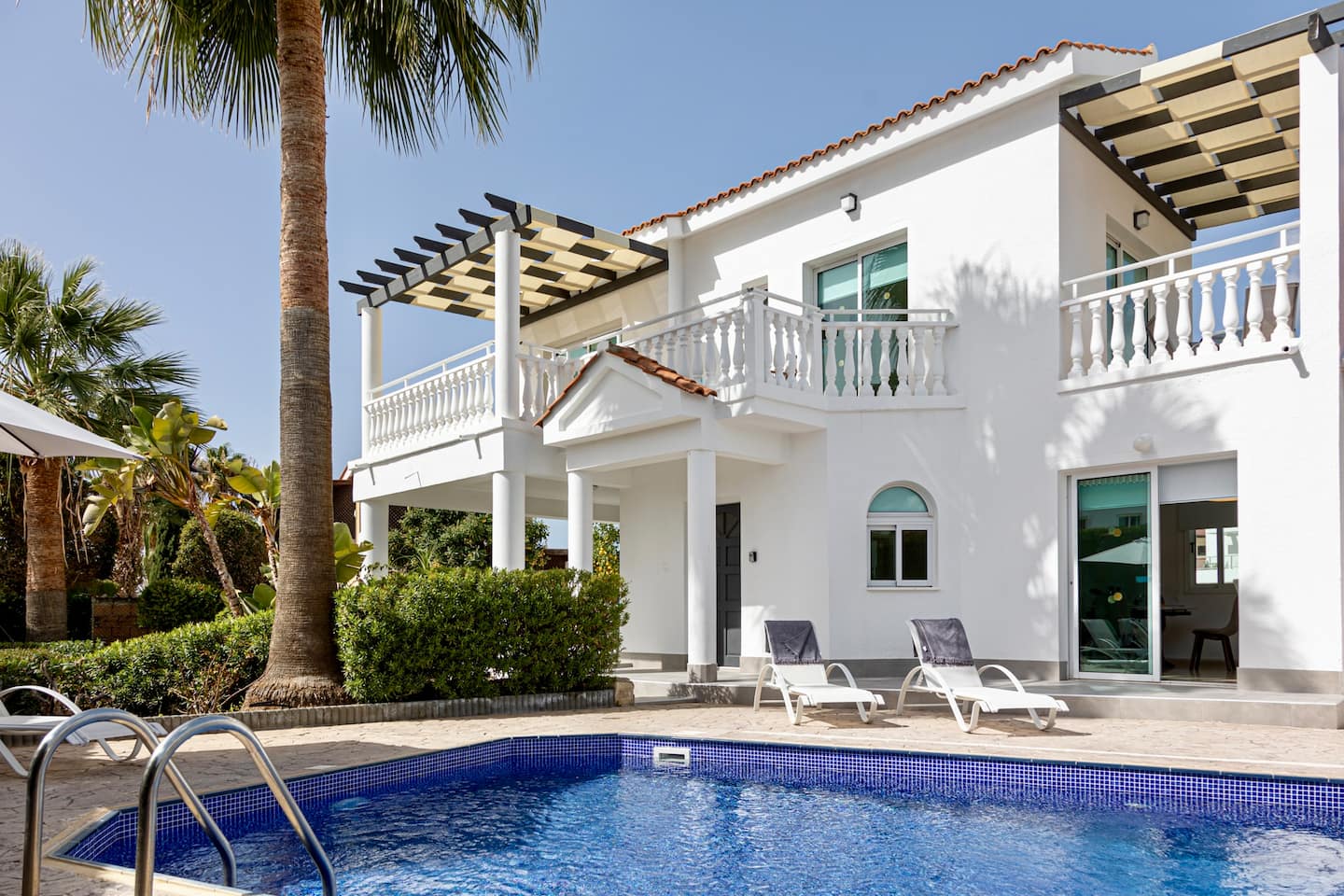 Whitewashed two-storey villa with balcony pergolas behind a blue mosaic swimming pool and sun loungers