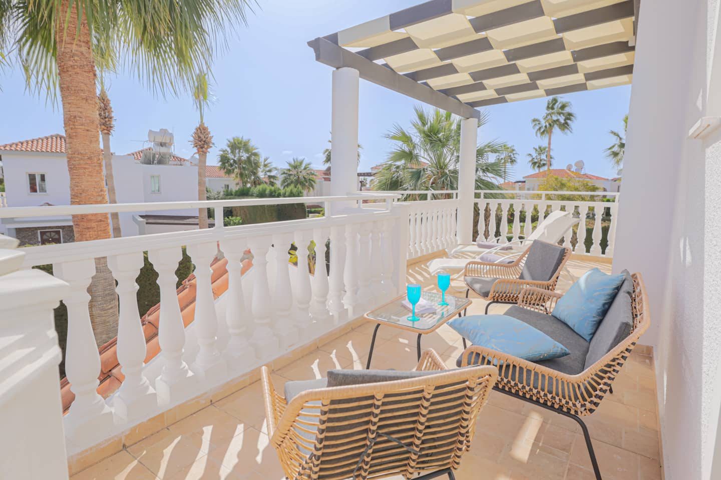 Rattan sofa and chairs on a pergola-shaded balcony above palms and tiled roofs